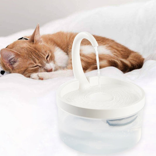 Cat drinking from white fountain – Cat resting beside an automatic pet water fountain on a white blanket.