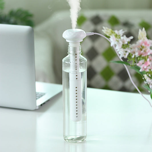 A white USB stick humidifier, inserted into a clear water bottle and emitting mist, sits on a white desk beside a blurred laptop.