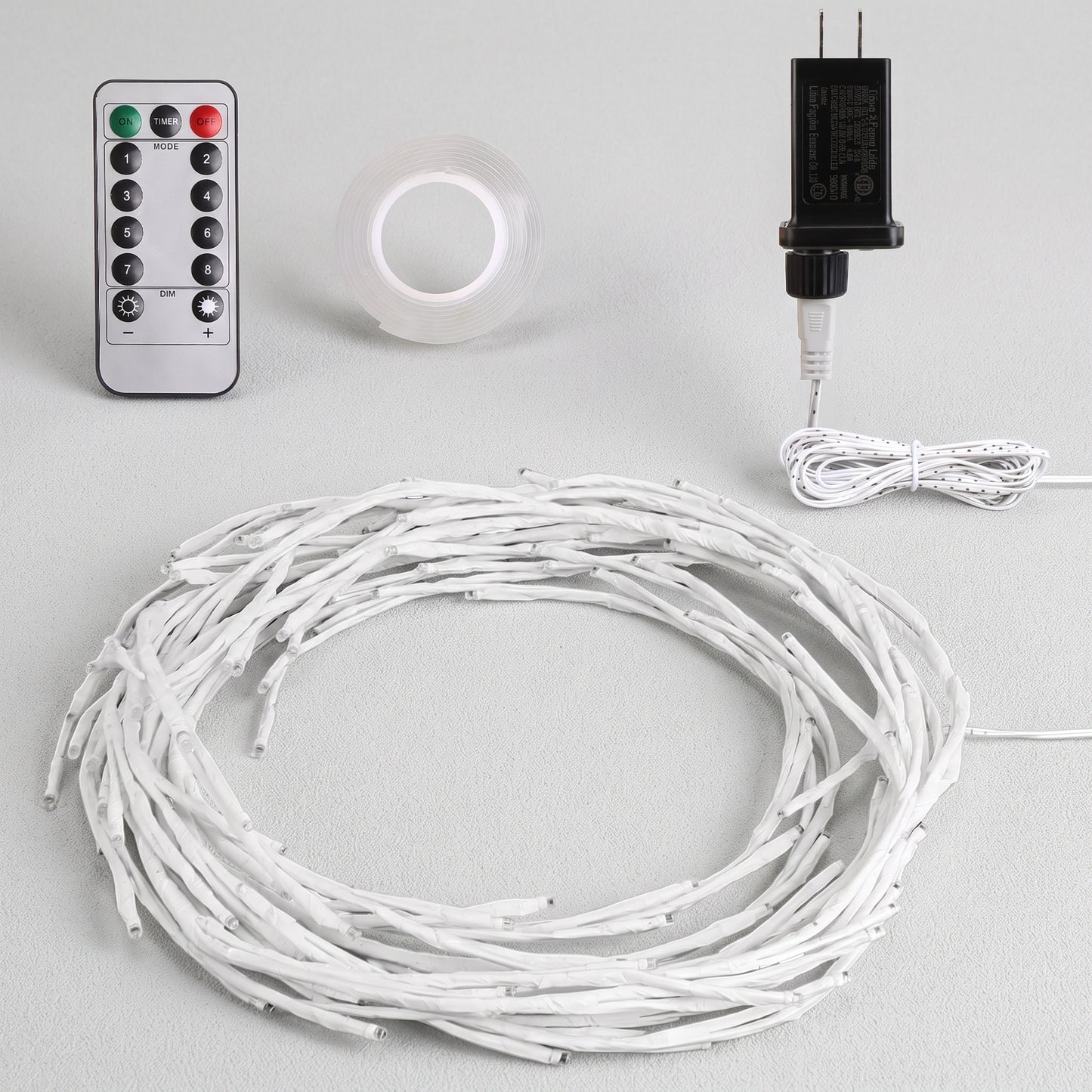 White LED string lights coiled in a large circle, shown with a silver remote control, a roll of clear adhesive tape, and a black power adapter on a light gray background.