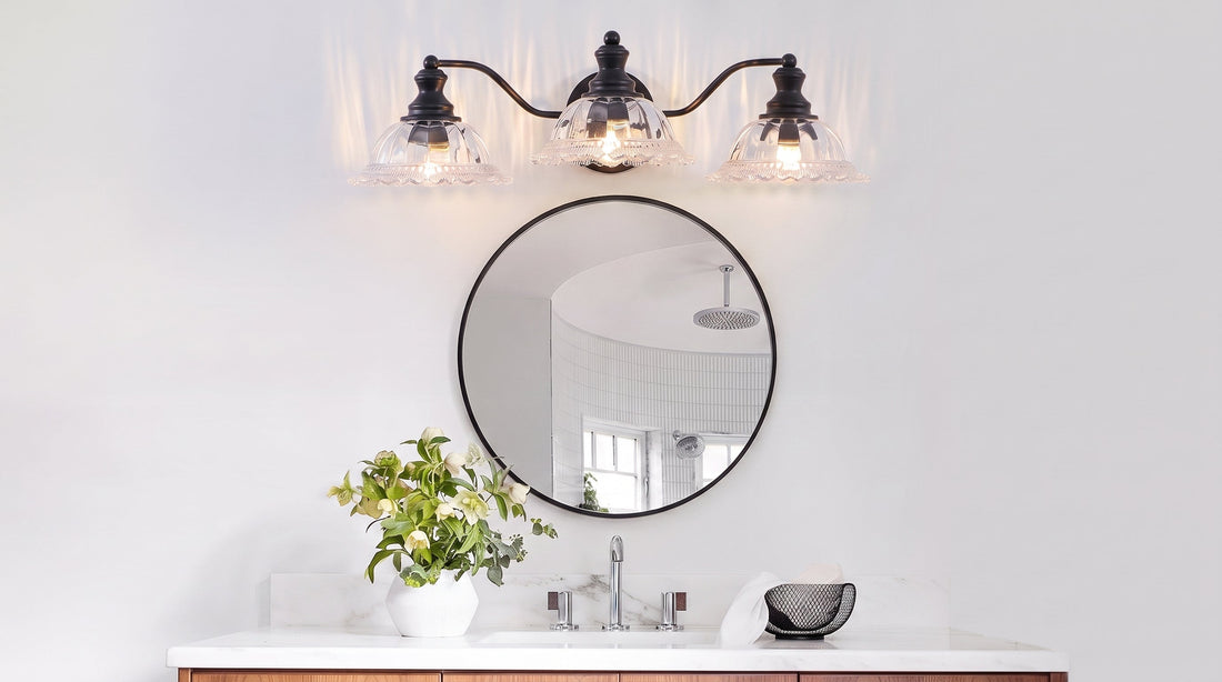 A stylish bathroom features a 3-light black vanity sconce above a round mirror, illuminating a white marble vanity with a vase of flowers.