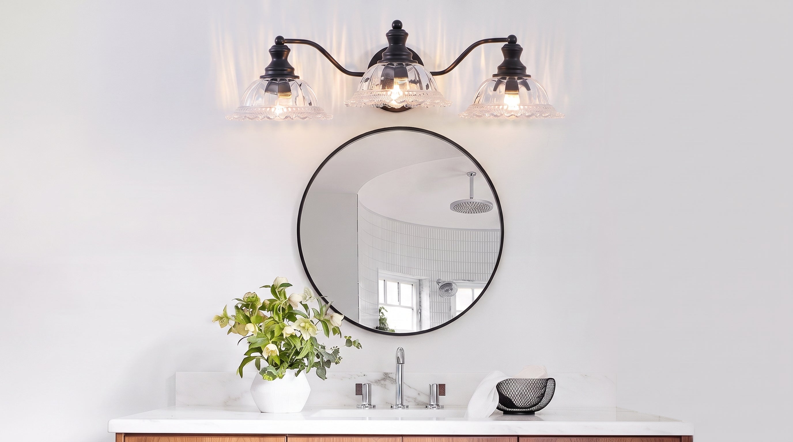 A stylish bathroom features a 3-light black vanity sconce above a round mirror, illuminating a white marble vanity with a vase of flowers.