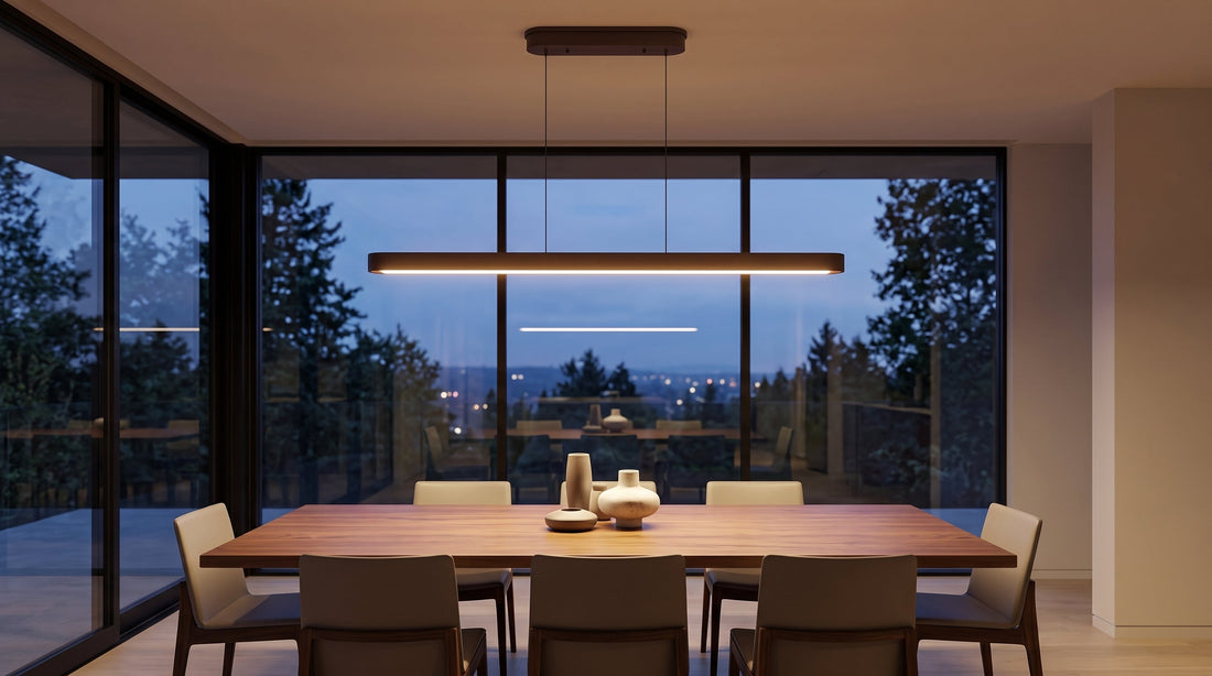 A sleek linear LED pendant light illuminates a modern dining room at dusk. The warm glow highlights a walnut table and chairs, with large windows overlooking a scenic forest and city lights.
