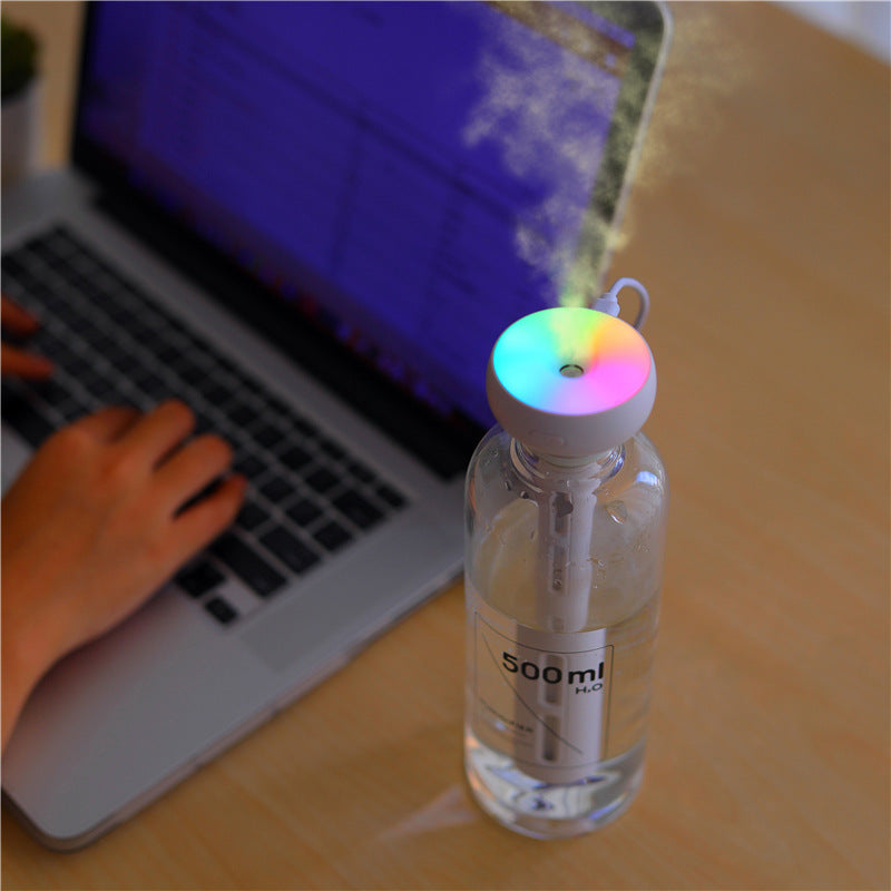 A ZAINZIO Mini USB Stick Humidifier in a "500ml H₂O" bottle, emitting mist while its colorful LED top glows, next to a laptop where someone is typing.