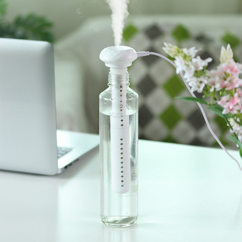 A white USB stick humidifier, inserted into a clear water bottle and emitting mist, sits on a white desk beside a blurred laptop.