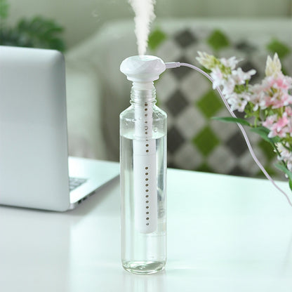 A white USB stick humidifier, inserted into a clear water bottle and emitting mist, sits on a white desk beside a blurred laptop.