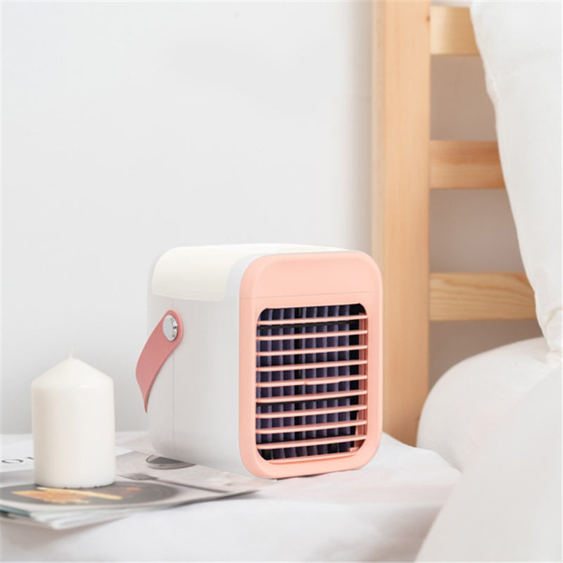 A compact white and pink portable misting cooler with a pink handle is shown on a nightstand. It sits next to a white candle and a magazine, emphasizing its small size.