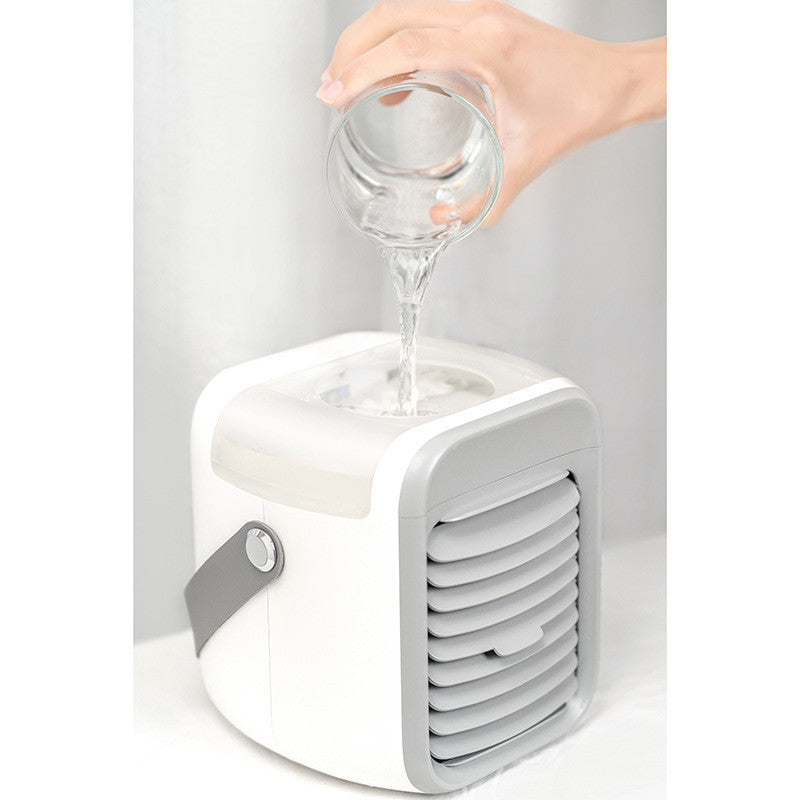 A hand pours water from a clear glass into the top of a compact white and gray Zainzio portable water cooling fan.