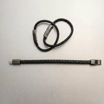 A black braided leather charging cable, displayed both as a stylish bracelet with a metallic clasp and straightened, revealing USB-A and USB-C connectors.