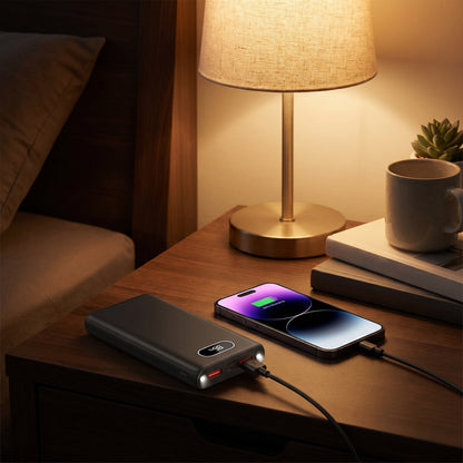 Power bank charging a smartphone on a bedside table with a lamp and mug in the background.