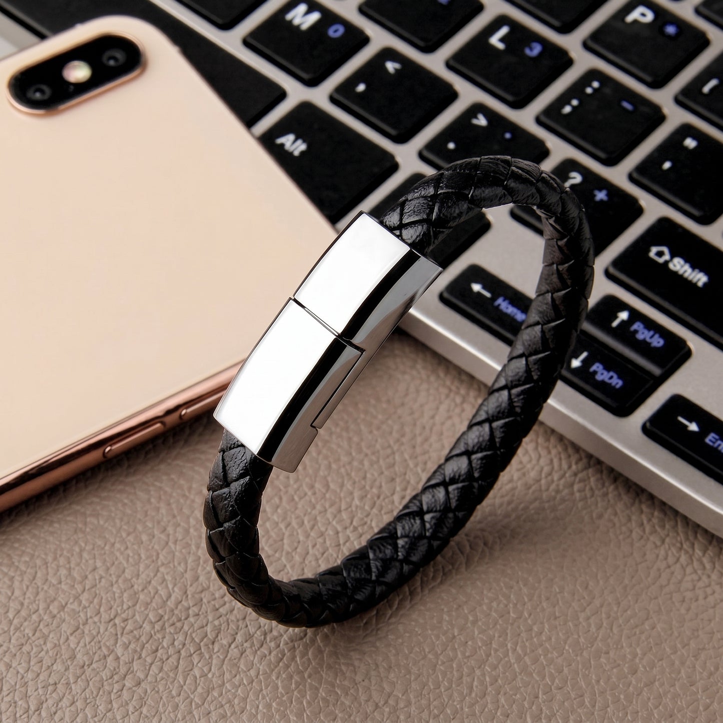 A black braided leather bracelet with a shiny silver USB-C clasp rests on a laptop keyboard near a gold smartphone.
