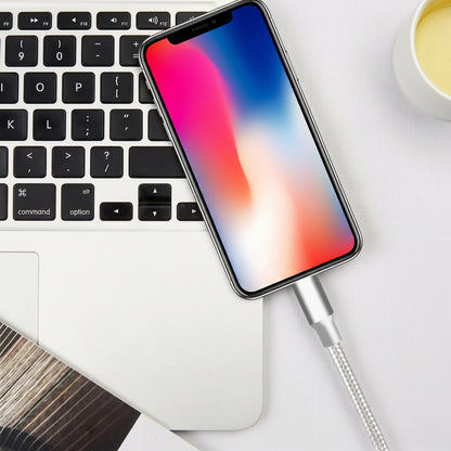 A silver braided charging cable connects an iPhone to a white MacBook, with a white mug in the background. It demonstrates the cable's use.