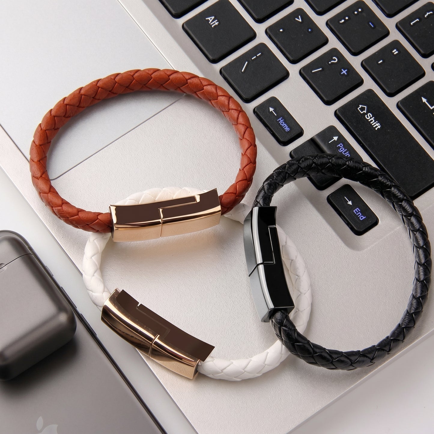 Three braided leather USB-C charging cable bracelets in brown, white, and black, resting on a laptop keyboard. They highlight the product's wearable design as power cables.