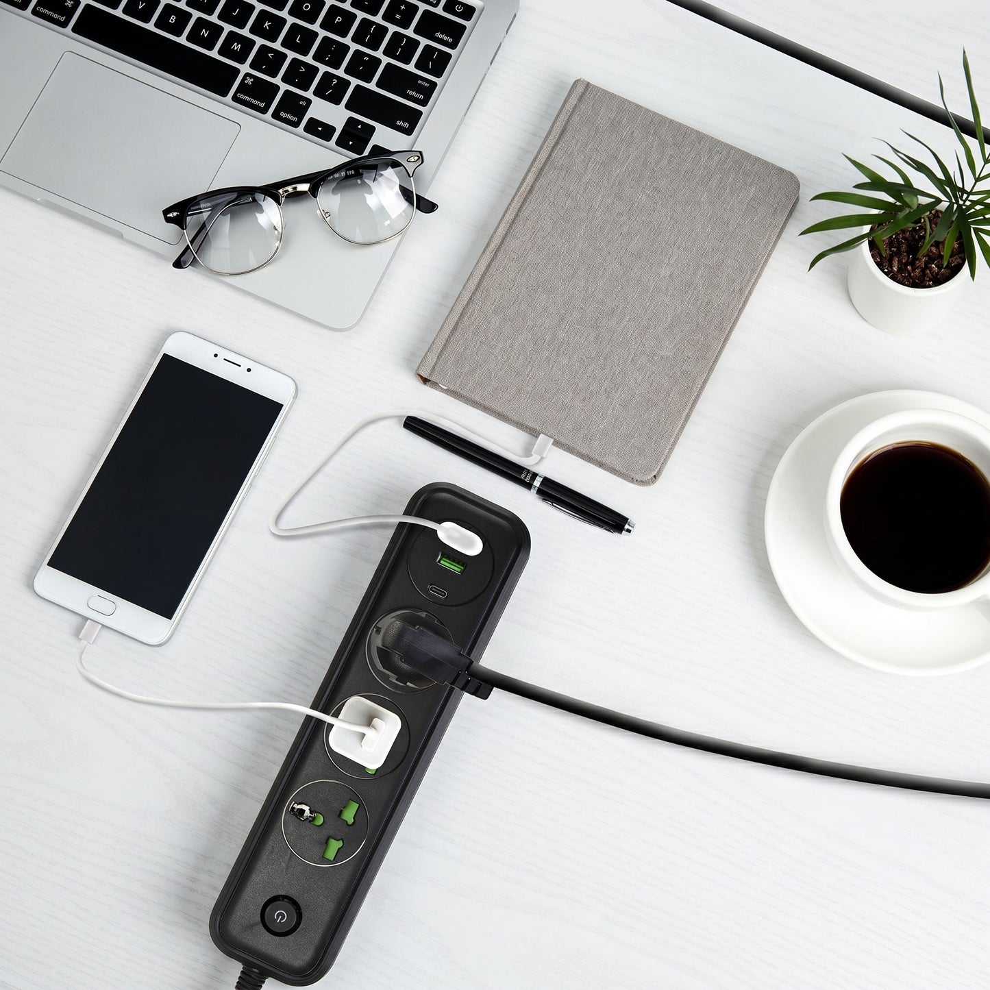 A flat lay shows a black power strip charging a smartphone and another device, alongside a laptop, notebook, and coffee on a white desk.