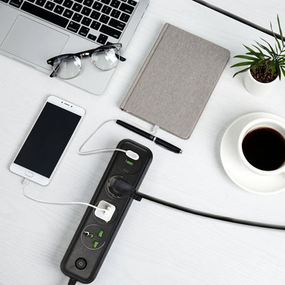 A flat lay shows a black power strip charging a smartphone and another device, alongside a laptop, notebook, and coffee on a white desk.