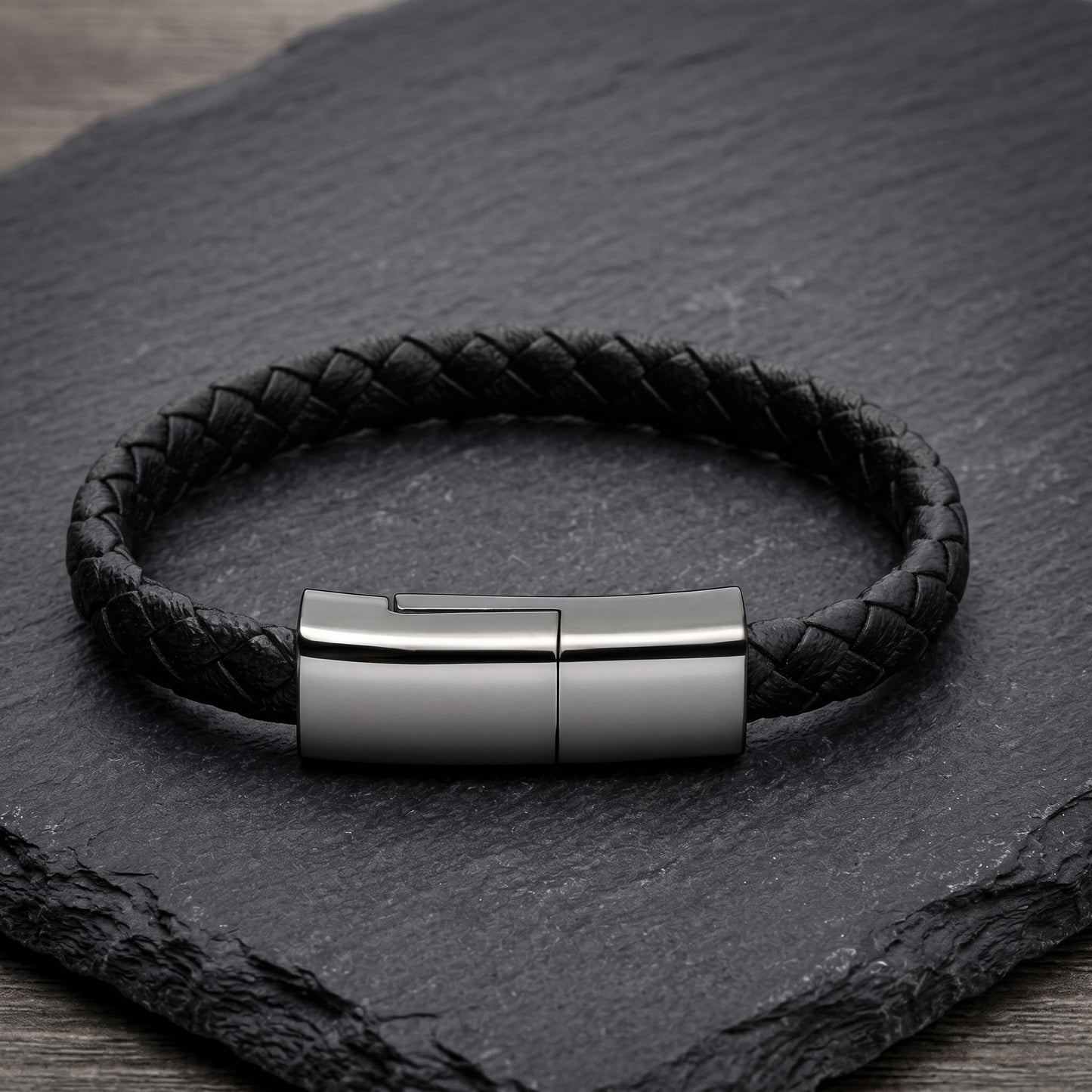 A black braided leather bracelet with a sleek, rectangular gunmetal clasp. This versatile accessory discretely functions as a USB-C charging cable.