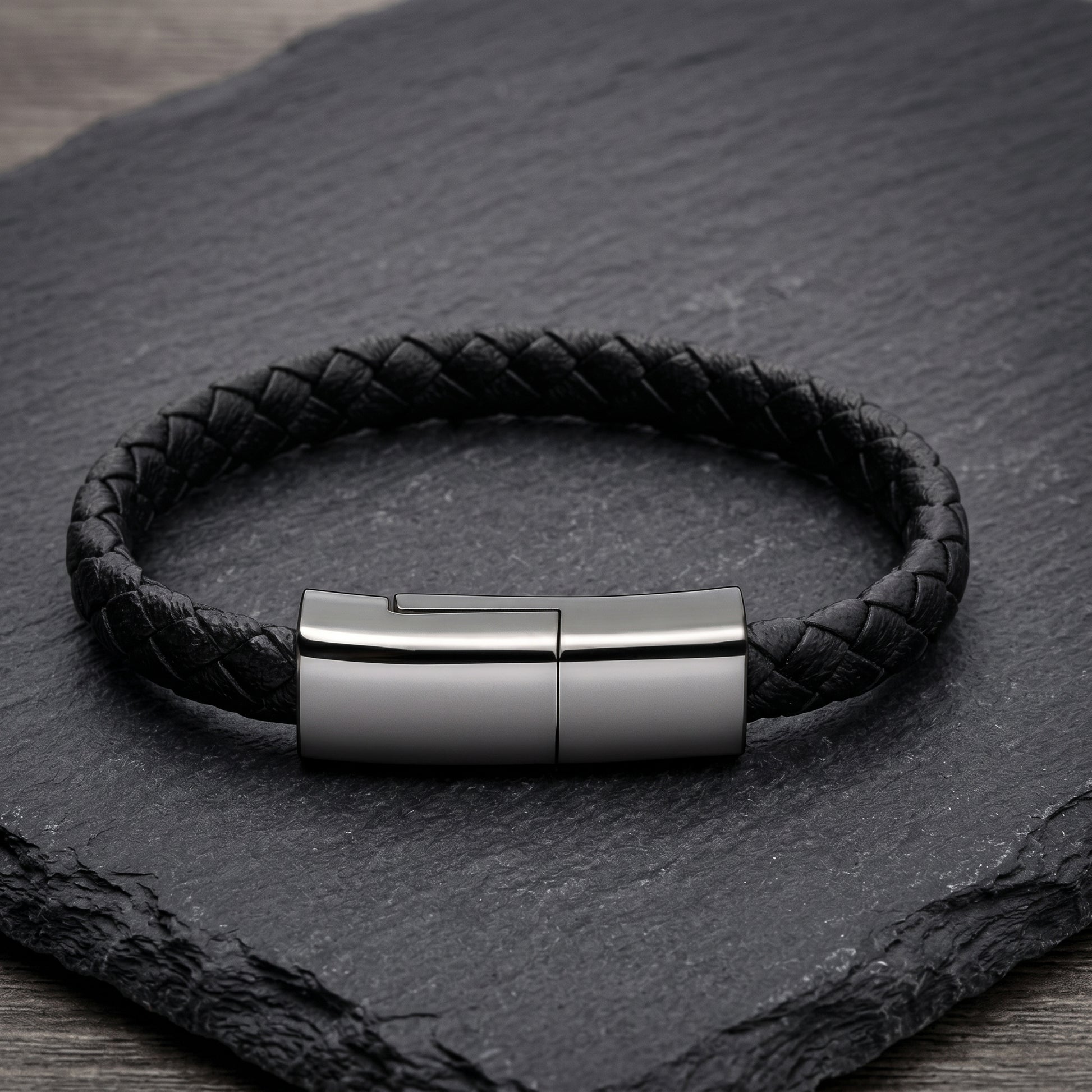 A black braided leather bracelet with a sleek, rectangular gunmetal clasp. This versatile accessory discretely functions as a USB-C charging cable.