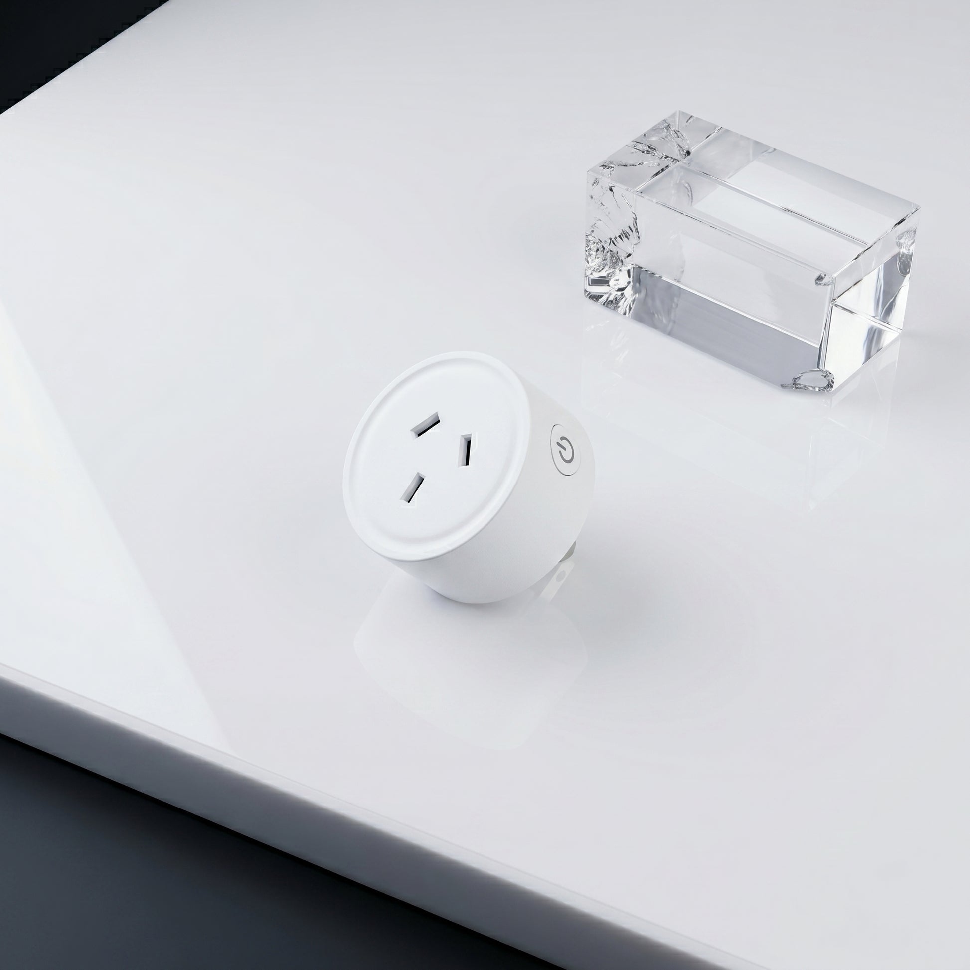 A white cylindrical smart plug and a clear block sit on a glossy white surface. The plug features three outlet holes and a power button with a power icon.