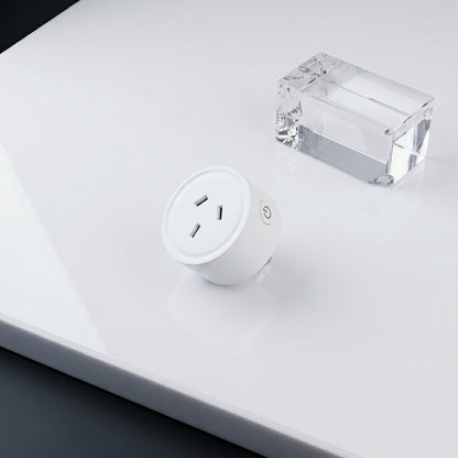 A white cylindrical smart plug and a clear block sit on a glossy white surface. The plug features three outlet holes and a power button with a power icon.