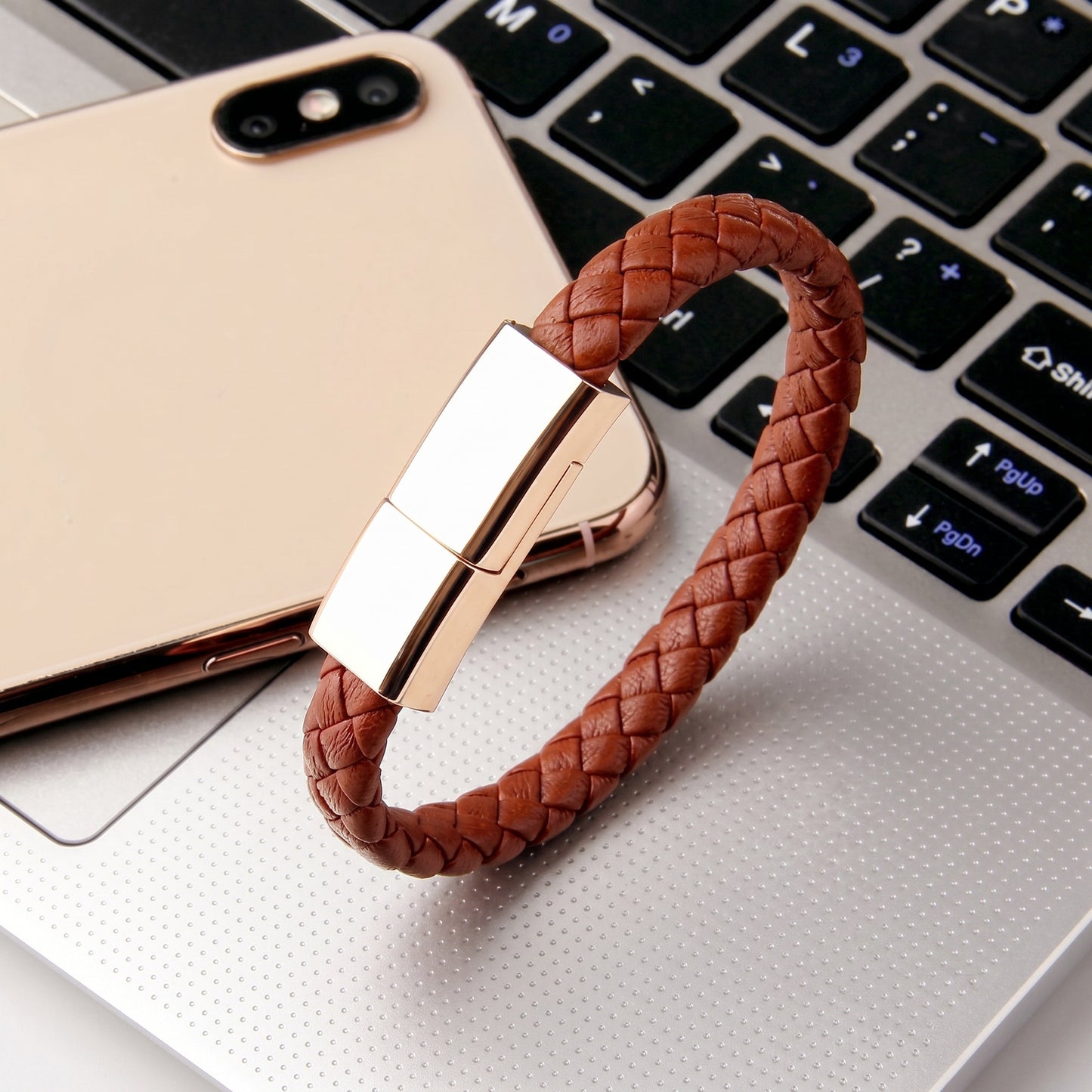 A brown braided leather bracelet, which doubles as a USB-C charging cable, rests on a laptop keyboard next to a gold smartphone.