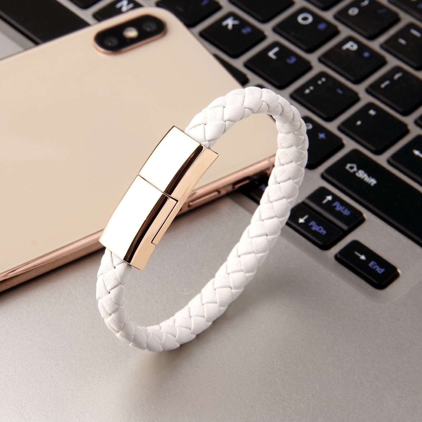 A white braided leather USB-C charging cable bracelet with a gold clasp, presented on a smartphone and laptop keyboard.
