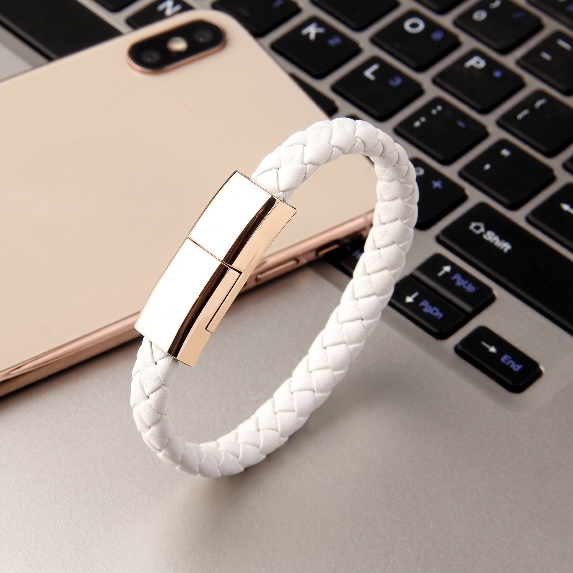 A white braided leather USB-C charging cable bracelet with a gold clasp, presented on a smartphone and laptop keyboard.