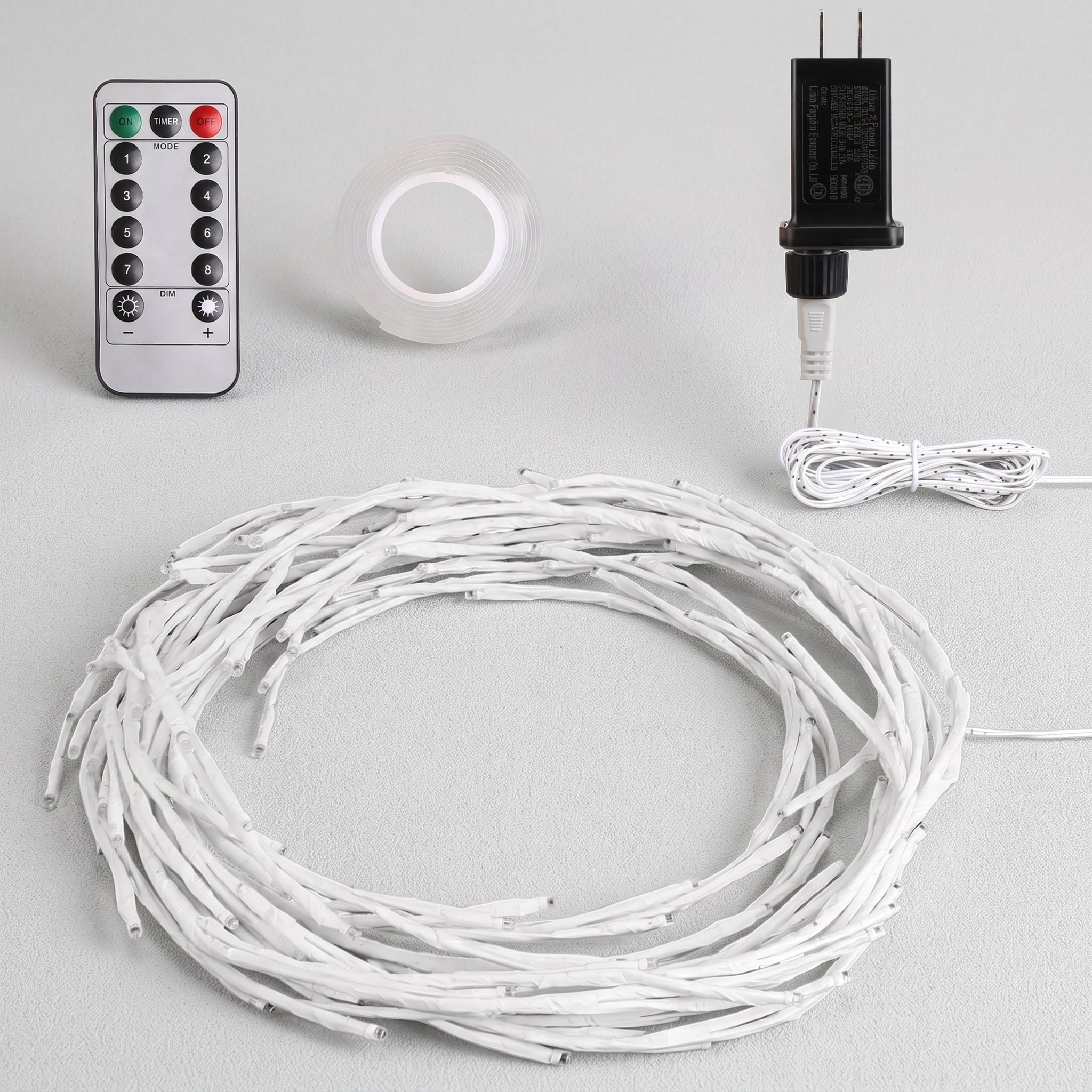 White LED string lights coiled in a large circle, shown with a silver remote control, a roll of clear adhesive tape, and a black power adapter on a light gray background.
