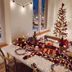 A cozy dining table decorated for Christmas with white tablecloths, patterned plates, wine bottles, and warm string lights, alongside a small decorated tree and a window view of a snowy outdoor scene at night.