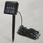 Solar-powered garden string lights with a black rectangular solar panel on a stake and a coiled bundle of green wire lights on a gray textured background.