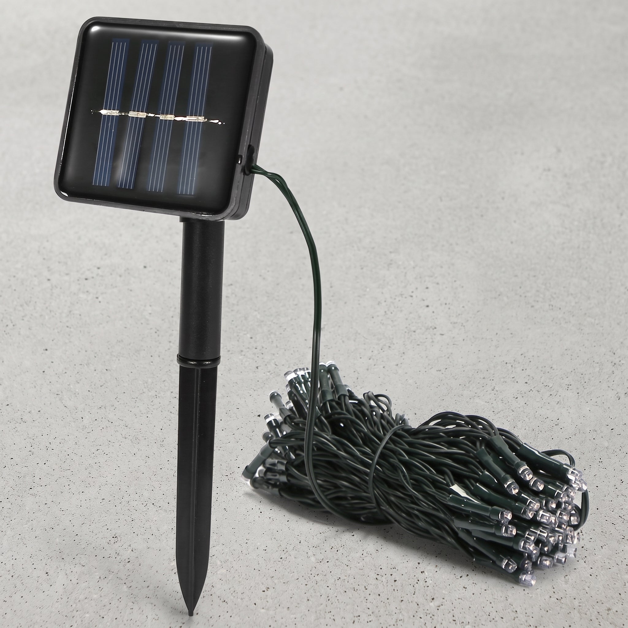 Solar-powered garden string lights with a black rectangular solar panel on a stake and a coiled bundle of green wire lights on a gray textured background.