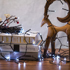 White fairy string lights draped over stacked books and a wooden deer figurine on a tabletop against a neutral wall.