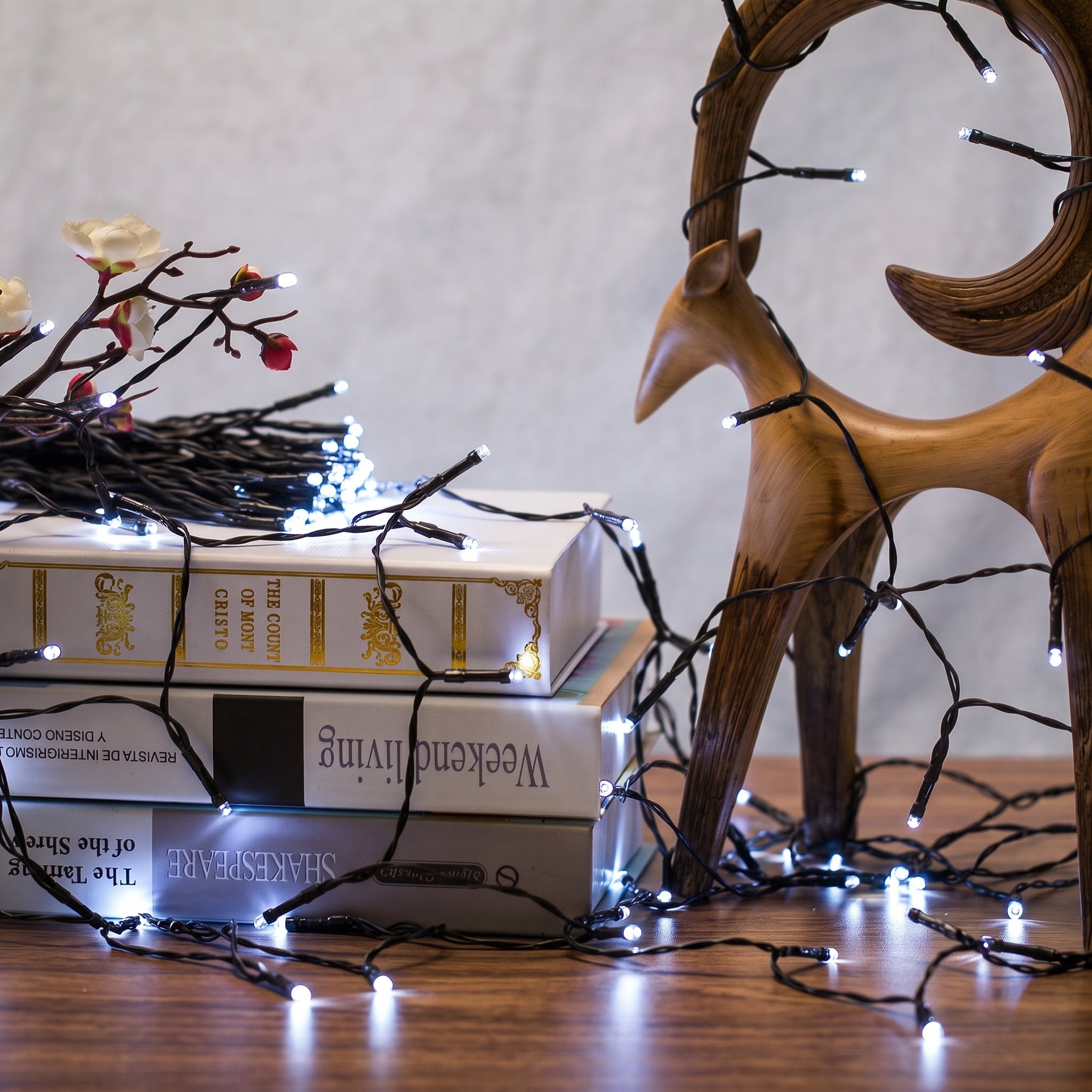 White fairy string lights draped over stacked books and a wooden deer figurine on a tabletop against a neutral wall.