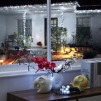 Decorative string lights with small white bulbs draped over a house porch at night, with red and white flowers and decorative vases in the foreground on a windowsill.