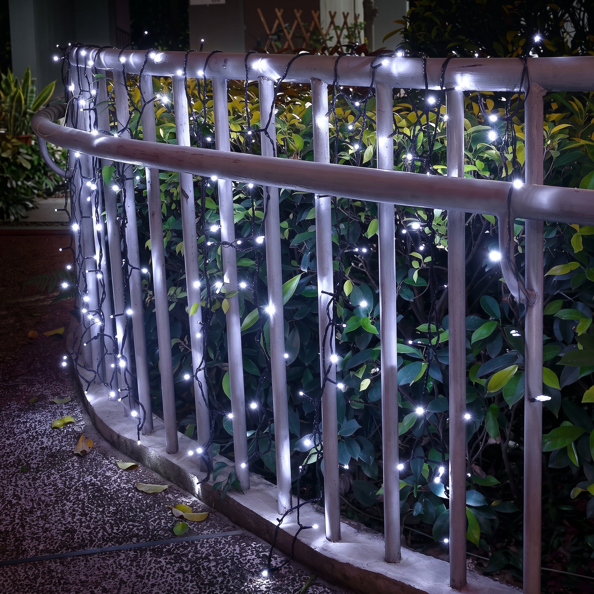 White LED string lights draped over a metal garden railing at night, casting a soft glow against green foliage in the background.