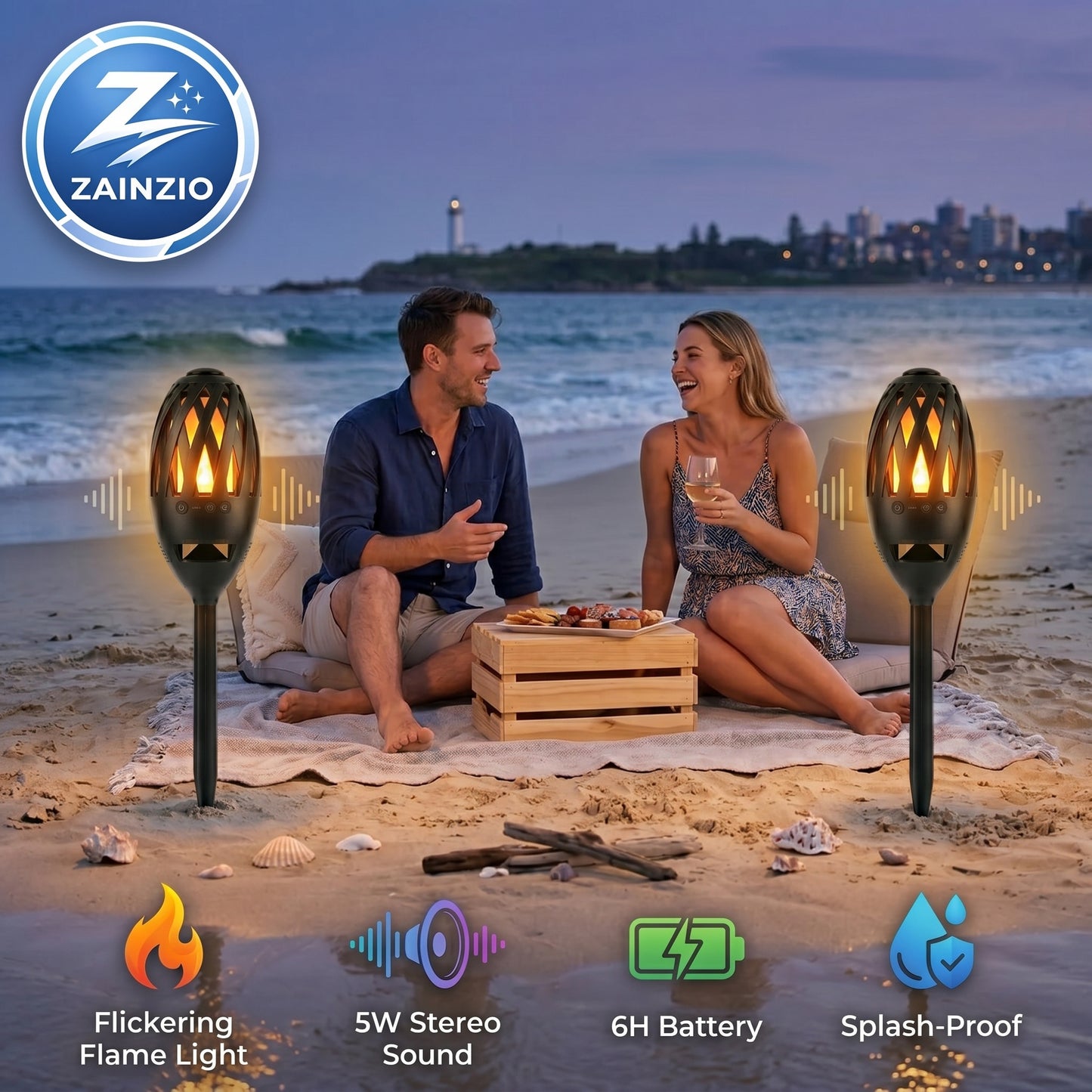 A couple enjoys a beach picnic at dusk, illuminated by two ZAINZIO flame speaker torches. Features include: Flickering Flame Light, 5W Stereo Sound, 6H Battery, Splash-Proof.