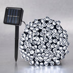 A solar-powered LED light string with a black stake-mounted solar panel on the left and a dense cluster of clear dome-shaped LED bulbs on the right, set against a neutral gray background.