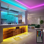 A modern bar and lounge illuminated by vibrant RGB LED strip lights in blue, pink, and yellow. A remote control, labeled "V+ V- OFF ON R G B W", is in the foreground.