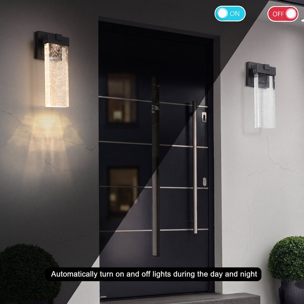 A pair of modern rectangular outdoor wall sconces mounted on either side of a black front door. The sconces have textured clear glass shades with black metal frames and are shown illuminated, casting warm light downward.
