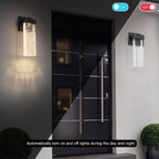 A pair of modern rectangular outdoor wall sconces mounted on either side of a black front door. The sconces have textured clear glass shades with black metal frames and are shown illuminated, casting warm light downward.