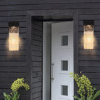 Two modern rectangular outdoor wall sconces with textured glass emit a warm glow on either side of a white front door set within a dark charcoal wood-clad building.