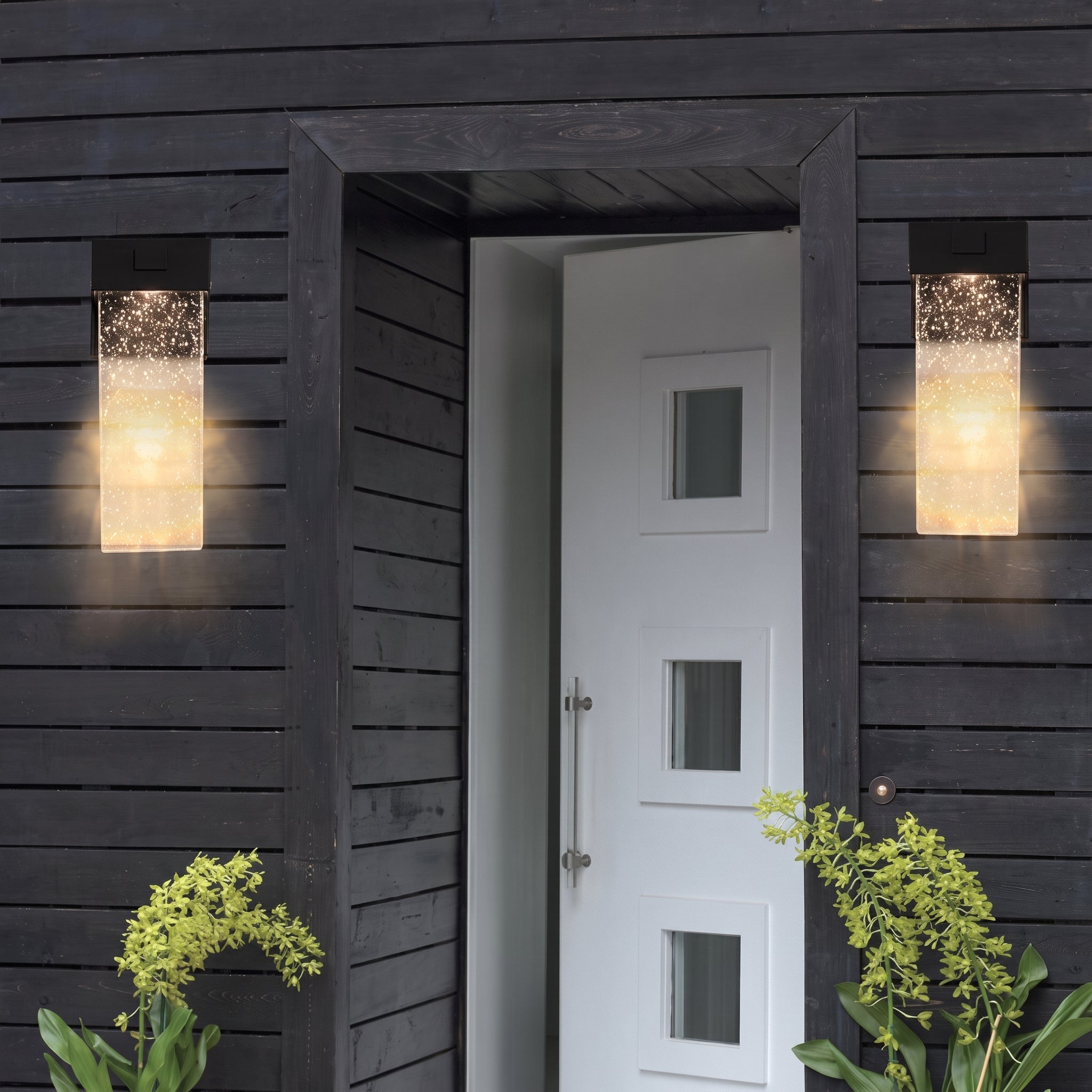 Two modern rectangular outdoor wall sconces with textured glass emit a warm glow on either side of a white front door set within a dark charcoal wood-clad building.