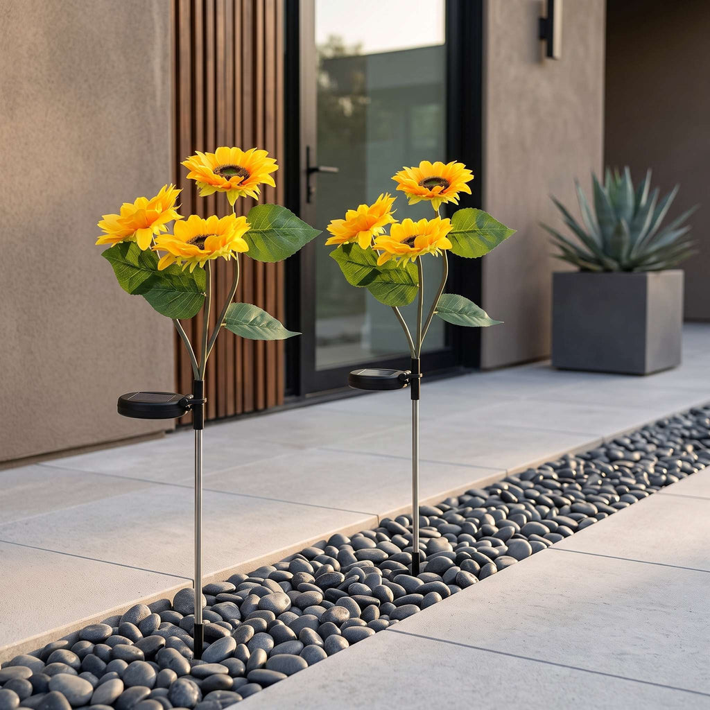 Two solar-powered sunflower garden lights with bright yellow petals and green leaves, planted in a stone garden bed beside a modern patio walkway.