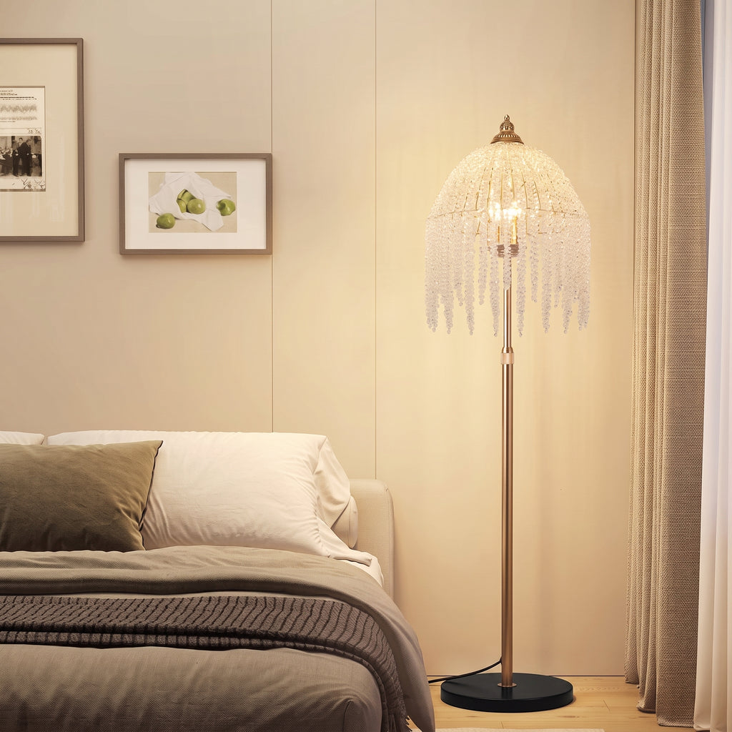 A modern floor lamp with a bronze-finished stand and a large, fringed bell-shaped shade illuminated with warm light, placed in a bedroom next to a bed.
