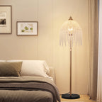 A modern floor lamp with a bronze-finished stand and a large, fringed bell-shaped shade illuminated with warm light, placed in a bedroom next to a bed.