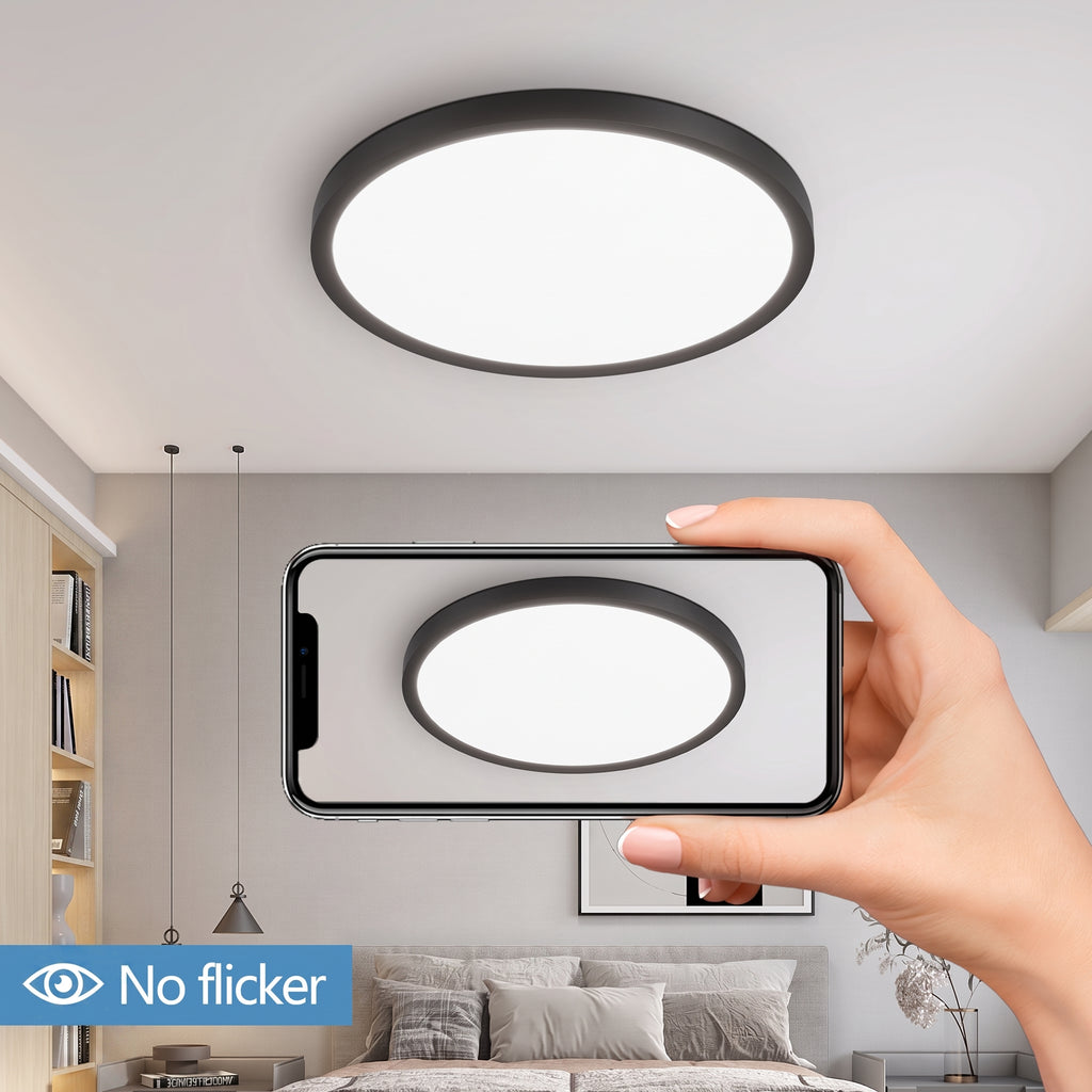 A round ceiling light with a black frame and white illuminated diffuser shown installed on a ceiling, with a hand holding a phone in front of it to demonstrate a close-up view. The room background includes a bed, wall art, pendant lights, and a bookshelf.