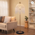 A floor lamp with a gold-toned metal stand and a dome-shaped shade covered in hanging clear crystal-like beads, shown in a styled living room beside a beige sofa and shelving.