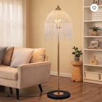 A floor lamp with a gold-toned metal stand and a dome-shaped shade covered in hanging clear crystal-like beads, shown in a styled living room beside a beige sofa and shelving.