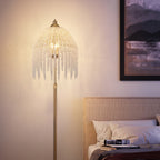 A bohemian-style floor lamp with a gold-finished pole and a dome-shaped shade covered in hanging clear crystal beads, glowing with warm light in a bedroom setting next to a bed.