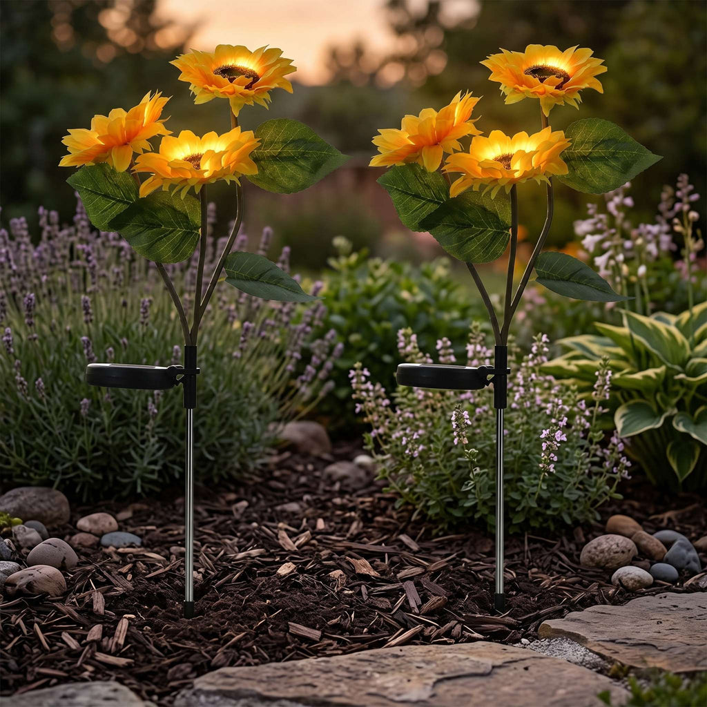 Two solar-powered sunflower garden lights with glowing yellow petals and green leaves, mounted on metal stakes in a mulched garden bed with rocks and plants.