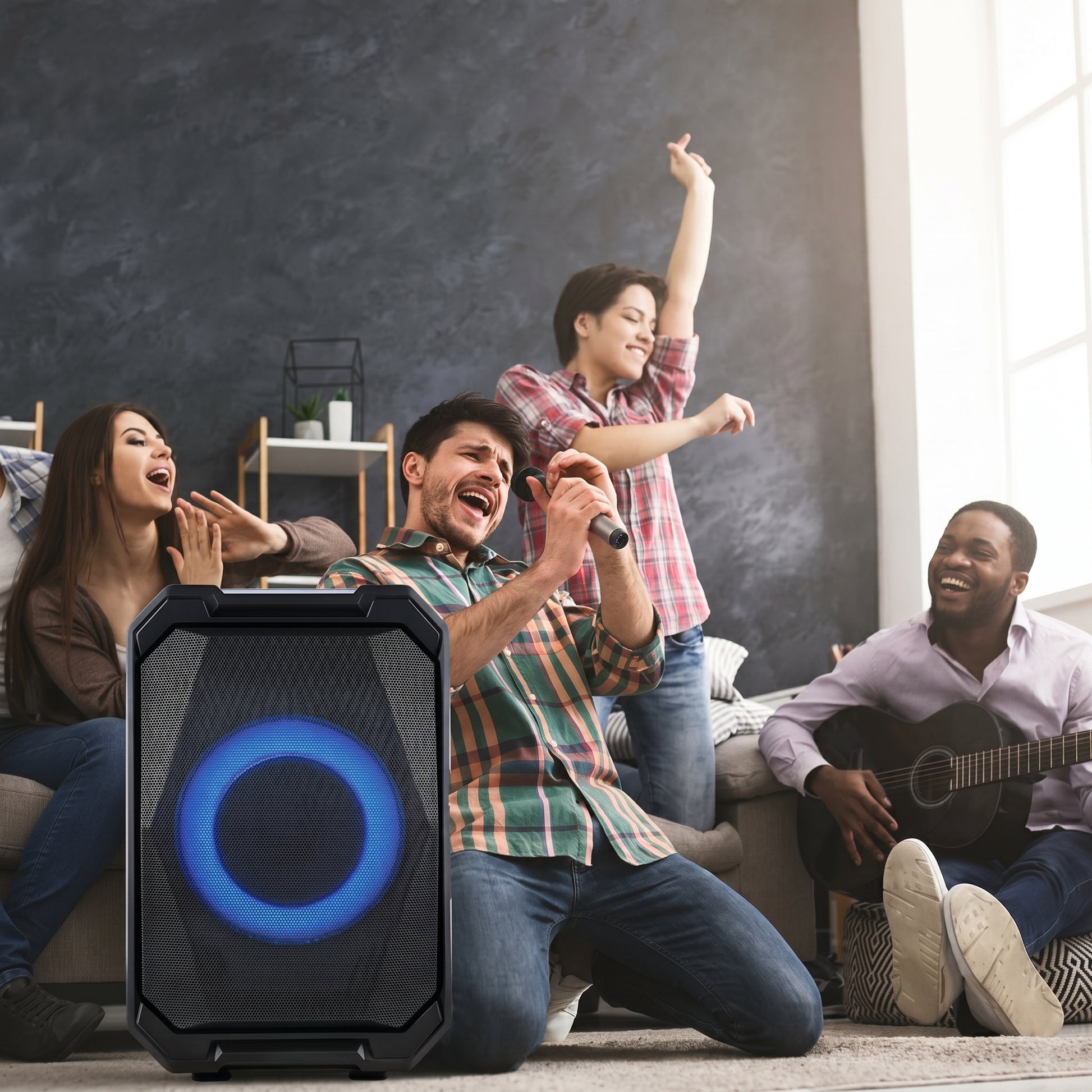 A black portable party speaker with a glowing blue light in the foreground. Four diverse friends are singing, dancing, and playing guitar in the background, enjoying the music.