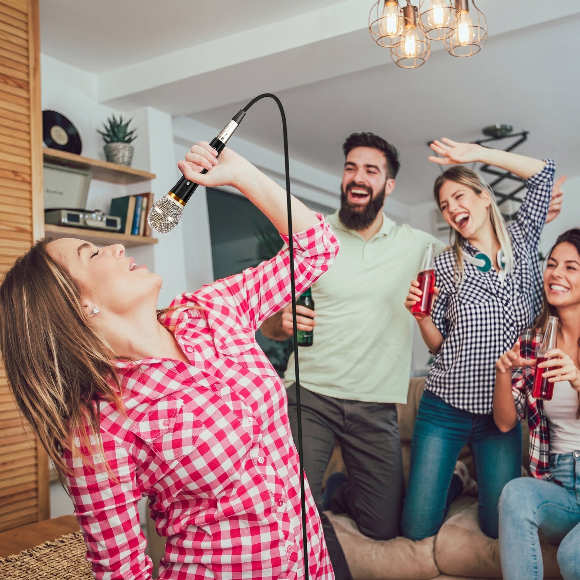 A woman sings enthusiastically into a black and silver wired microphone at a lively home karaoke party, with friends smiling and dancing in the background.